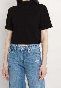 Svart kortärmad t-shirt i bomull med hög halsringning och rullade korta ärmar; kombinerad med blå, slitna vida jeans med fickdetalj.