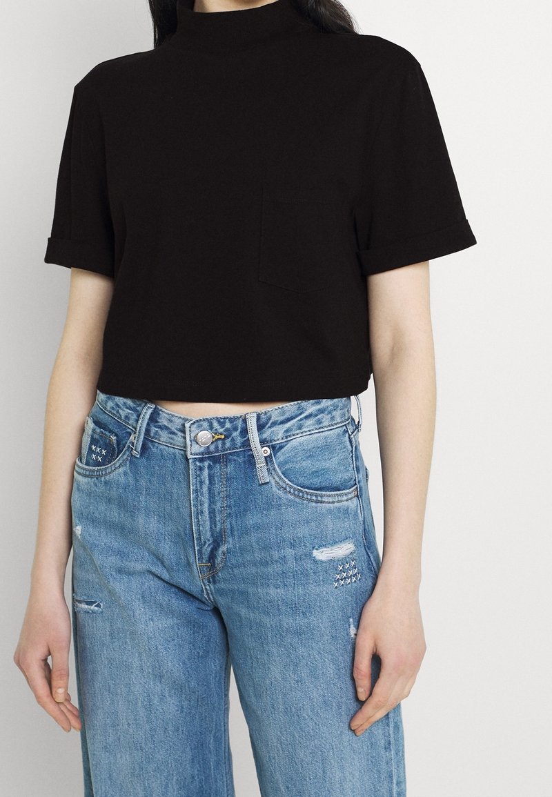 Svart kortärmad t-shirt i bomull med hög halsringning och rullade korta ärmar; kombinerad med blå, slitna vida jeans med fickdetalj.