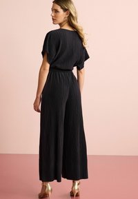 Svart, plisserad jumpsuit med vida ben och korta ärmar. Har en cinchad midja och en texturerad yta. Bärs med guldiga klacksandaler.
