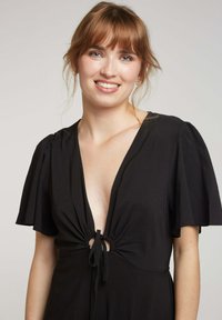 Robe noire avec de courtes manches flottantes, décolleté en V profond et détail noué à la taille. Fabriquée à partir d'un tissu doux et léger avec une finition texturée.