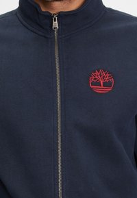 Námořnická zipová bunda vyrobená z měkké látky, s vyšitým červeným logo stromu na levém hrudníku a plnohodnotným stříbrným zipem.