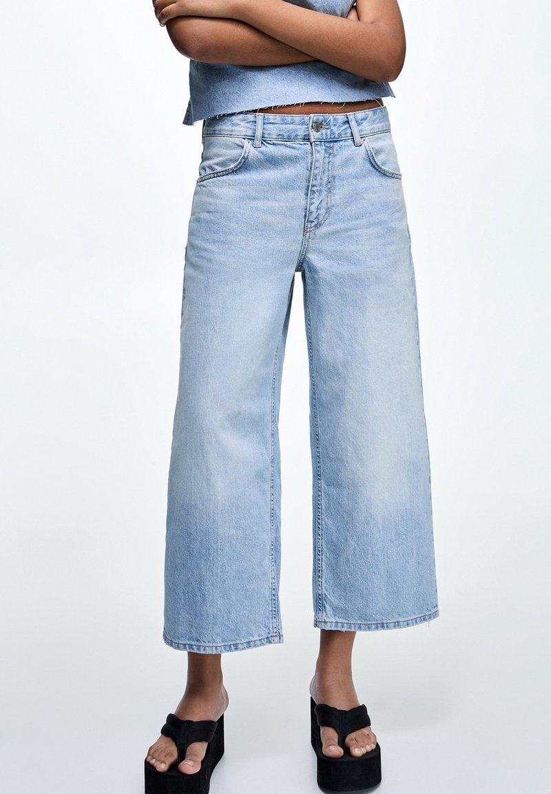 Vaqueros de pierna ancha de color azul claro, confeccionados en denim, con cintura alta, cinco bolsillos y detalles de dobladillo deshilachado. Se llevan con sandalias negras de plataforma.