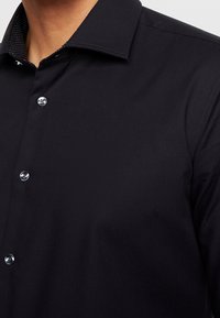  närbild av en person som bär en svart skjorta med knappar och krage, där den svarta innerfoder syns med vita prickar.