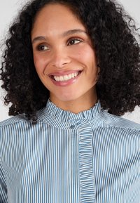 Blauw en wit gestreept shirt met een ruches kraag en een voorpas. De stof lijkt glad met een lichte glans, wat het patroon benadrukt.