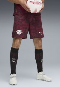 Pantalones cortos de fútbol con un patrón en rojo y negro, con un logo, acompañados de calcetas negras con acentos blancos y un balón de fútbol blanco con un logo.