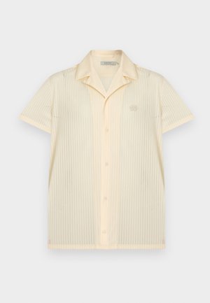 Młody mężczyzna z kręconymi czarnymi włosami, ubrany w beżową krótką koszulę na krótki rękaw rozpiętą z przodu, białą koszulkę, niebieskie jeansowe szorty, białe tenisówki i skarpetki.