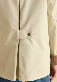 Jachetă ușoară bej, cu deschidere la spate, având închidere cu nasture și manșete cu nervuri. Textură netedă și design simplu.