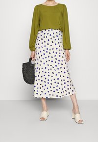 Blouse verte à manches longues au-dessus d'une jupe crème avec un motif tacheté bleu et noir, associée à des sandales à talons blocs beiges et un sac en osier.