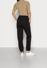 Pantalones de chándal negros con cinturilla y puños elásticos. Combinados con una camiseta ajustada beige y zapatillas blancas. Se muestra la vista trasera.