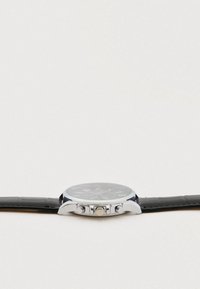 Silbernes Uhrengehäuse mit schwarzem Zifferblatt und drei Tasten an der Seite, befestigt an einem schwarzen Lederarmband, flach liegend auf einer weißen Oberfläche.