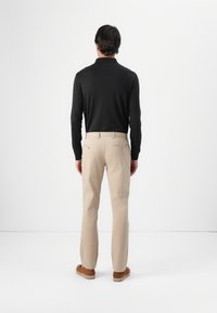 Zwarte lange mouwen top, beige broek met dubbele achterzakken en bruine instapschoenen. Minimalistisch ontwerp tegen een effen achtergrond.
