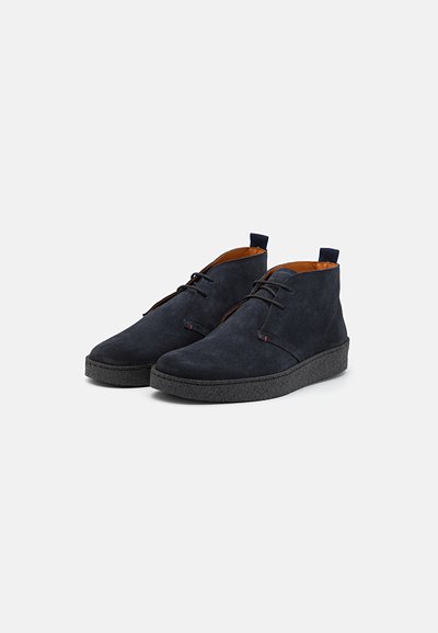 Botas de ante azul marino hasta el tobillo con una suela negra texturizada, que presentan un diseño de cordones y un forro marrón claro en contraste. Incluyen una pestaña en la parte posterior.