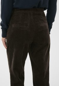 Pantalon en velours côtelé marron avec des rayures verticales, des poches arrière et une taille haute. Tissu texturé avec une apparence douce et côtelée.
