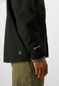 Jachetă impermeabilă neagră, cu branding Gore-Tex, textură netedă și căptușeală din fleece gri în contrast la măneci.
