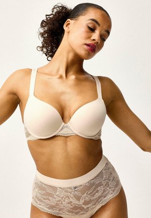 Frau posiert in beigem, gepolstertem BH und hoch tailliertem Spitzenunterwäsche vor einem schlichten hellen Hintergrund.