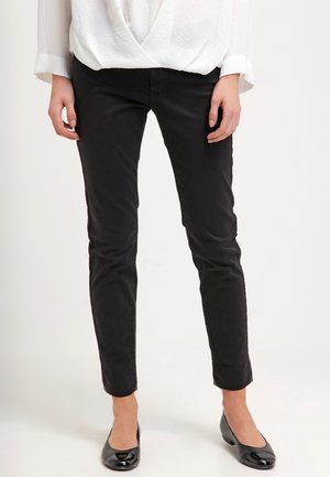 Persoon die strakke zwarte broek, een witte top met lange mouwen en glanzende zwarte schoenen met lage hak draagt, staande tegen een effen achtergrond.