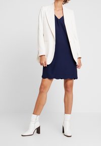 Vit skräddarsydd kavaj över en marinblå klänning med vågade kanter, matchad med vita ankelboots. Släta tyger, strukturerad design och kontrasterande färger.