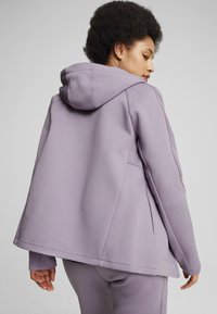 Sudadera morada con un ajuste relajado, textura suave y bolsillo tipo canguro. Presenta una capucha amplia y detalles de costura en contraste a lo largo de las costuras.