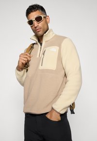 Beige fleece genser med kvart glidelås, en glidelås frontlomme og en kontrasterende kremfarget panel. Har teksturert stoff og en logo.