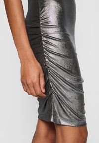 Gros plan sur une robe argentée scintillante froncée sur la hanche et la cuisse d'une personne, avec sa main gauche posée calmement à côté du tissu.