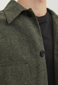 Giacca in misto lana verde scuro con colletto alla francese, chiusura con un bottone nero e tasca frontale; indossata sopra una camicia nera.