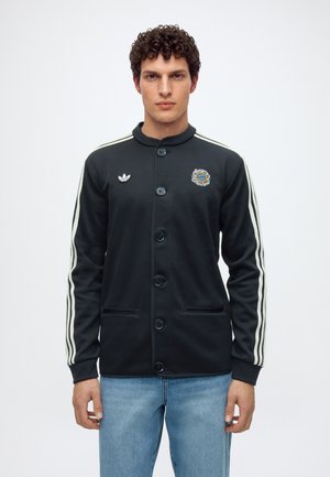 Млад мъж с къдрави коси, облечен в черно яке на Adidas с копчета и бели ивици по ръкавите, и сини дънки, стоящ на бял фон.