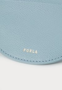 Pelle azzurro chiaro con superficie testurizzata, caratterizzata da un marchio "FURLA" in oro su un font minuscolo. Bordo arrotondato e cuciture precise.