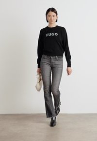 Zwarte trui met "HUGO" logo, grijze flared jeans met een ruwe zoom, zwarte enkellaarsjes, een witte tas vasthoudend, staand op een lichte vloer.