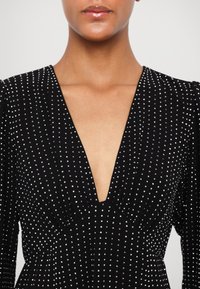 Femme portant une robe noire avec un profond décolleté en V et un motif à pois blancs, montrant le cou et le haut de la poitrine sur un fond uni.