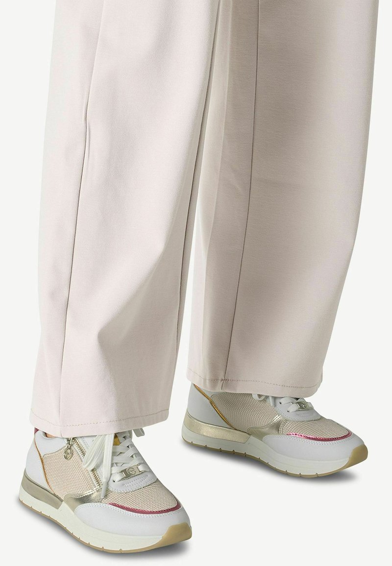 Weiße Sneaker mit Mesh und goldenen Akzenten, versehen mit einem dezenten rosa Detail. Kombiniert mit lockeren, hellbeigen Hosen.