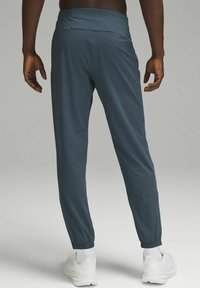 Herrarnas slim-fit joggers i en mörk teal-färg, tillverkade av lättviktsmaterial, med elastiska muddar och en slät textur. Vita sneakers bärs.