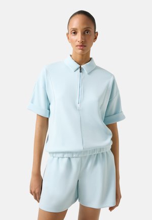 Kobieta z włosami zaczesanymi do tyłu, ubrana w jasnoniebieską koszulkę na suwak z krótkim rękawem i pasujące krótkie spodenki z elastycznym paskiem na jednolitym tle.