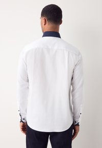 Camicia bianca a maniche lunghe con motivo a texture, colletto blu navy a contrasto e polsini con bottoni. Vestibilità sartoriale con orlo curvo.