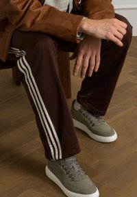 Bruine sportbroeken met witte strepen, gecombineerd met grijze schoenen met een gestructureerde bovenkant en witte zolen, zittend op een houten vloer.