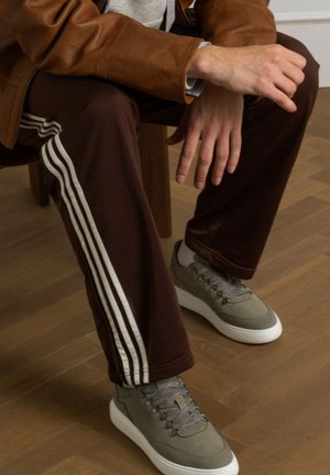 Bruine sportbroeken met witte strepen, gecombineerd met grijze schoenen met een gestructureerde bovenkant en witte zolen, zittend op een houten vloer.