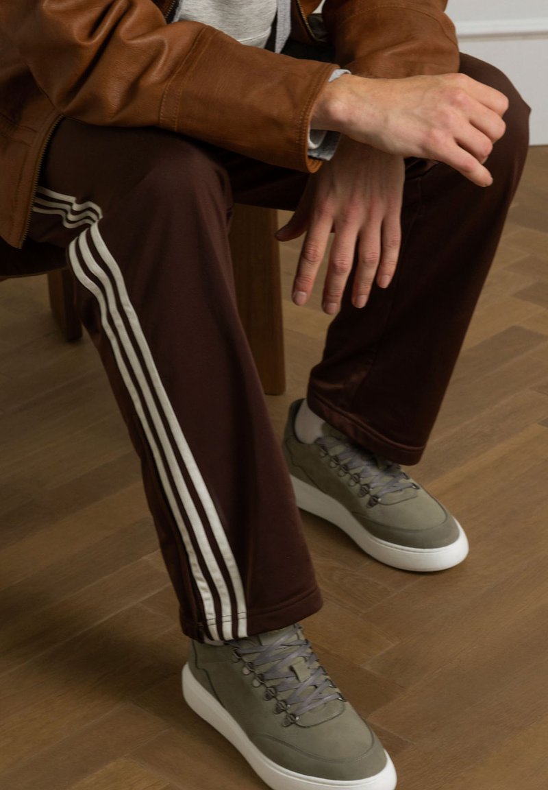 Bruine sportbroeken met witte strepen, gecombineerd met grijze schoenen met een gestructureerde bovenkant en witte zolen, zittend op een houten vloer.