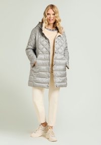 Silberner gesteppter Steppmantel mit Kapuze, mit Druckknopfverschluss und Seitentaschen, getragen über einem beigen Oberteil und hellen Hosen.