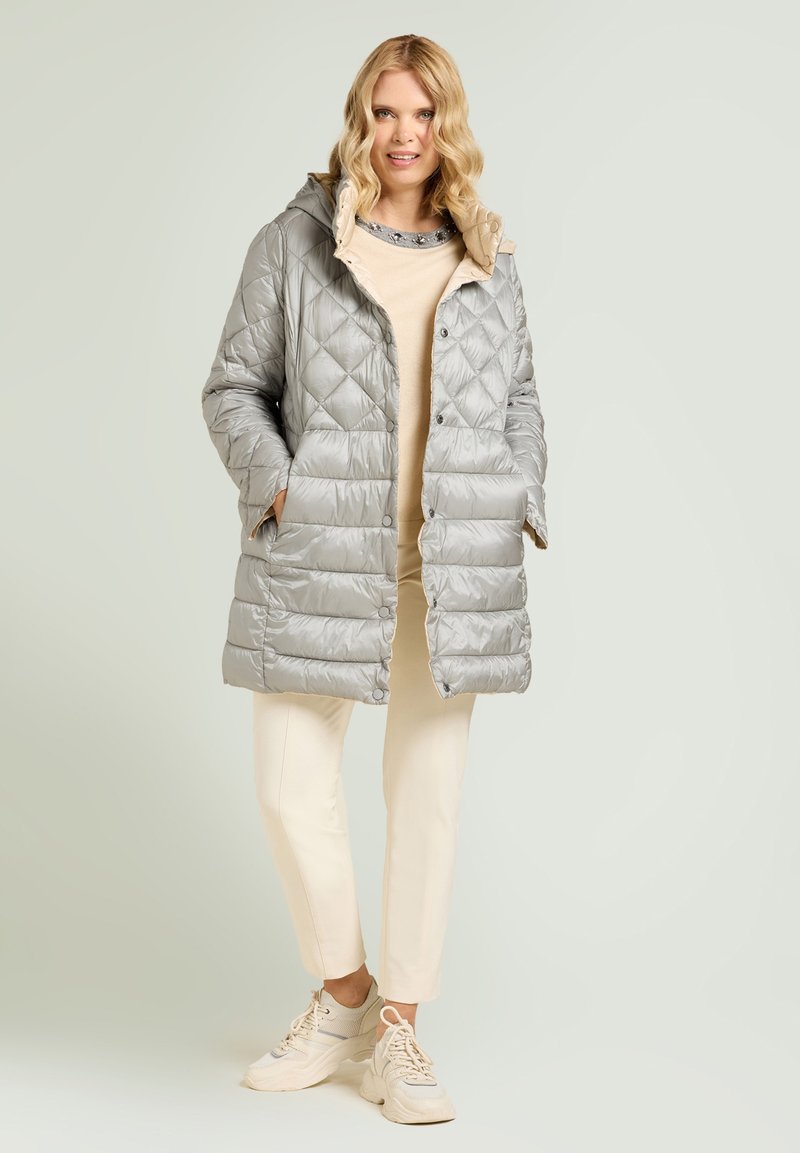 Silberner gesteppter Steppmantel mit Kapuze, mit Druckknopfverschluss und Seitentaschen, getragen über einem beigen Oberteil und hellen Hosen.