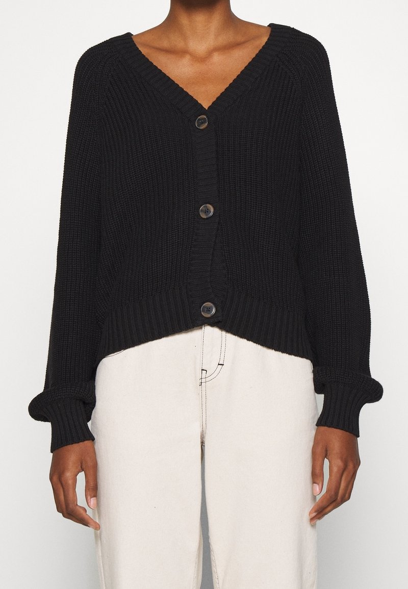Femme portant un cardigan en maille épaisse noir à col en V avec des boutons, associé à un pantalon taille haute de couleur ivoire.