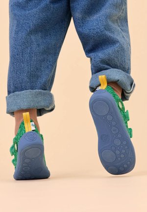 Bambino che cammina in avanti indossando scarpe di tessuto verde con suole blu strutturate e jeans blu risvoltati su uno sfondo beige.