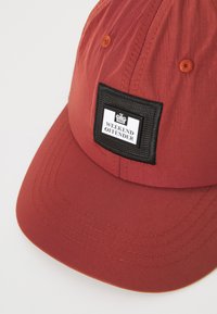 Rød cap laget av lett stoff med en buet skygge, med et svart rektangulært felt med teksten "WEEKEND OFFENDER" i hvitt.