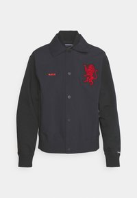 Veste marine avec broderie de lion rouge, fermeture à boutons et poignets côtelés. Présente un tissu lisse avec des manches texturées contrastantes.
