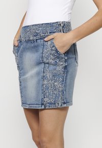Jupe en denim avec broderie florale sur le côté, de couleur bleu clair, design ajusté et poches sur le devant. Fermeture éclair à la taille.