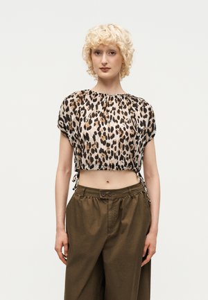 Frau mit kurzen lockigen blonden Haaren, die einen bauchfreien Leoparden-Print-Top und hoch taillierte, lockere braune Hosen trägt, steht vor einem schlichten Hintergrund.