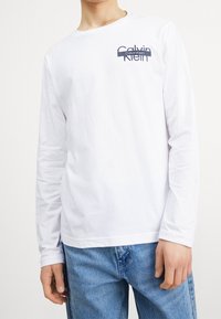Bílá dlouhá rukávová tričko s modrým logem Calvin Klein na levé straně hrudi. Tkanina působí hladce a lehce. Nosi se s modrými džíny.