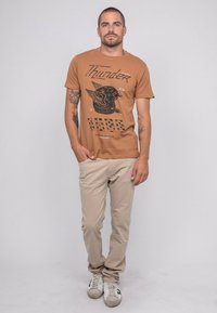 T-shirt graphique marron avec un motif imprimé noir représentant un oiseau et du texte. Associé à un pantalon beige clair et des baskets blanches avec des accents noirs.