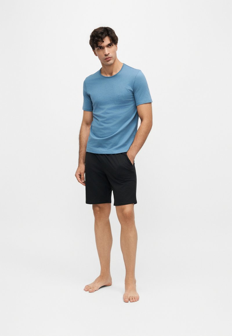 Homme debout pieds nus, portant un t-shirt bleu à manches courtes et un short noir jusqu'aux genoux sur un fond blanc uni.