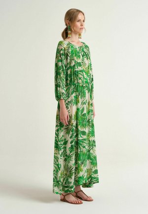 Femme portant une longue robe ample à imprimé feuilles tropicales vertes et blanches, manches longues, boucles d'oreilles à pompons colorés et sandales plates marron.
