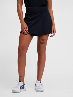 Femme portant un skort sportif noir et des baskets blanches, montrant un tatouage sur la cuisse d'une abeille avec "1982" et un tatouage à la cheville "SWS".