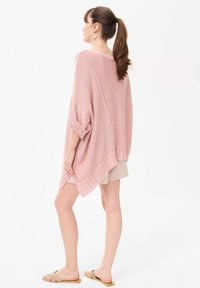 Lichtroze gebreide poncho met een losse pasvorm, opgerolde mouwen en een asymmetrische zoom. Textuurstof met open steeking op de achterkant.